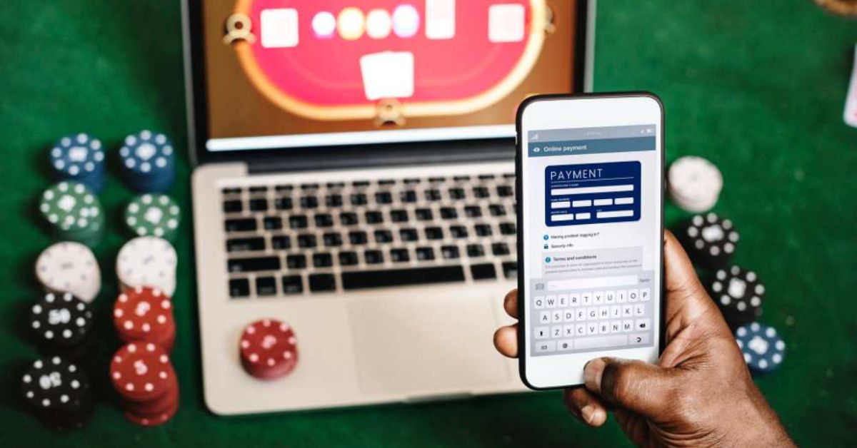 Person making an online payment on a smartphone with poker chips and a virtual casino table displayed on a laptop in the background.